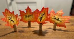 Colorful painted leaves from old book pages standing upright on corks, each with a name written in black, on a wooden table