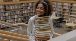 older Black teenage girl smiling with a stack of books in a library